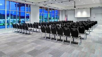Un salón de conferencias con filas de sillas, ventanas amplias y una pantalla de proyección.