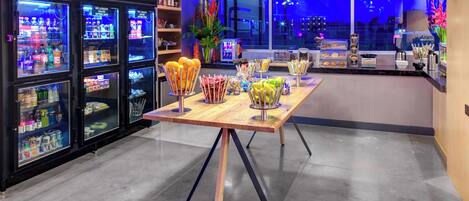A bar area with a long wooden table, various snack dispensers, and a refrigerator stocked with beverages.