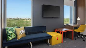 A modern hotel room with a blue sofa, a yellow ottoman, a desk, and a TV mounted on the wall.
