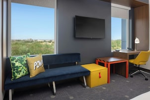 A modern hotel room with a blue sofa, a yellow ottoman, a desk, and a TV mounted on the wall.