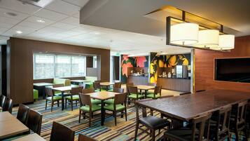 A hotel dining area with multiple tables and chairs, a large window, and a bar area with a colorful mural.