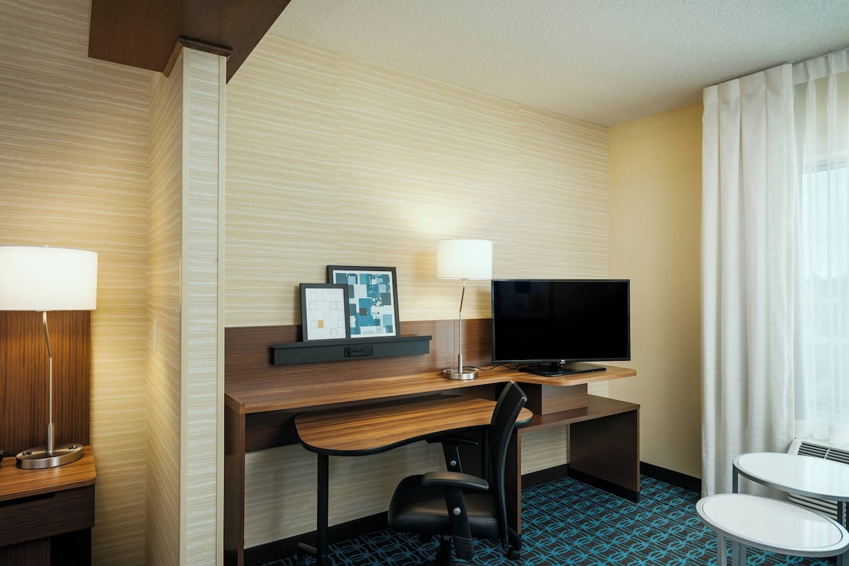 A hotel room with a wooden desk, a television, a chair, a small table, and a window with curtains.