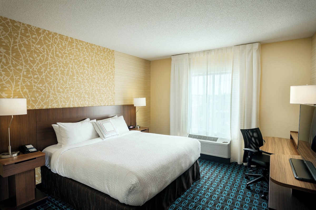 A hotel room with a large bed, two bedside tables with lamps, a desk with a chair, a television, and a window with curtains.