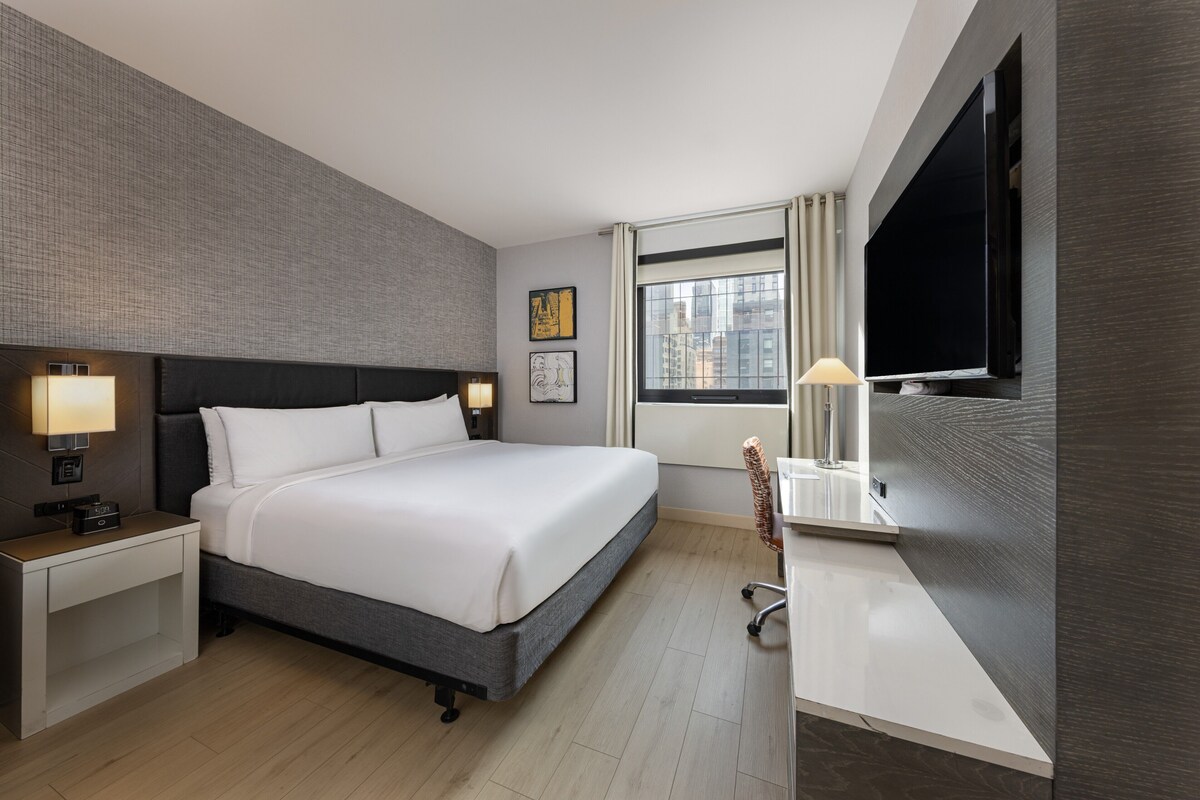 Ein modernes Hotelzimmer mit einem großen Bett, Nachttischen mit Lampen, einem Schreibtisch mit Stuhl, einem Flachbildfernseher und einem Fenster mit Vorhängen.