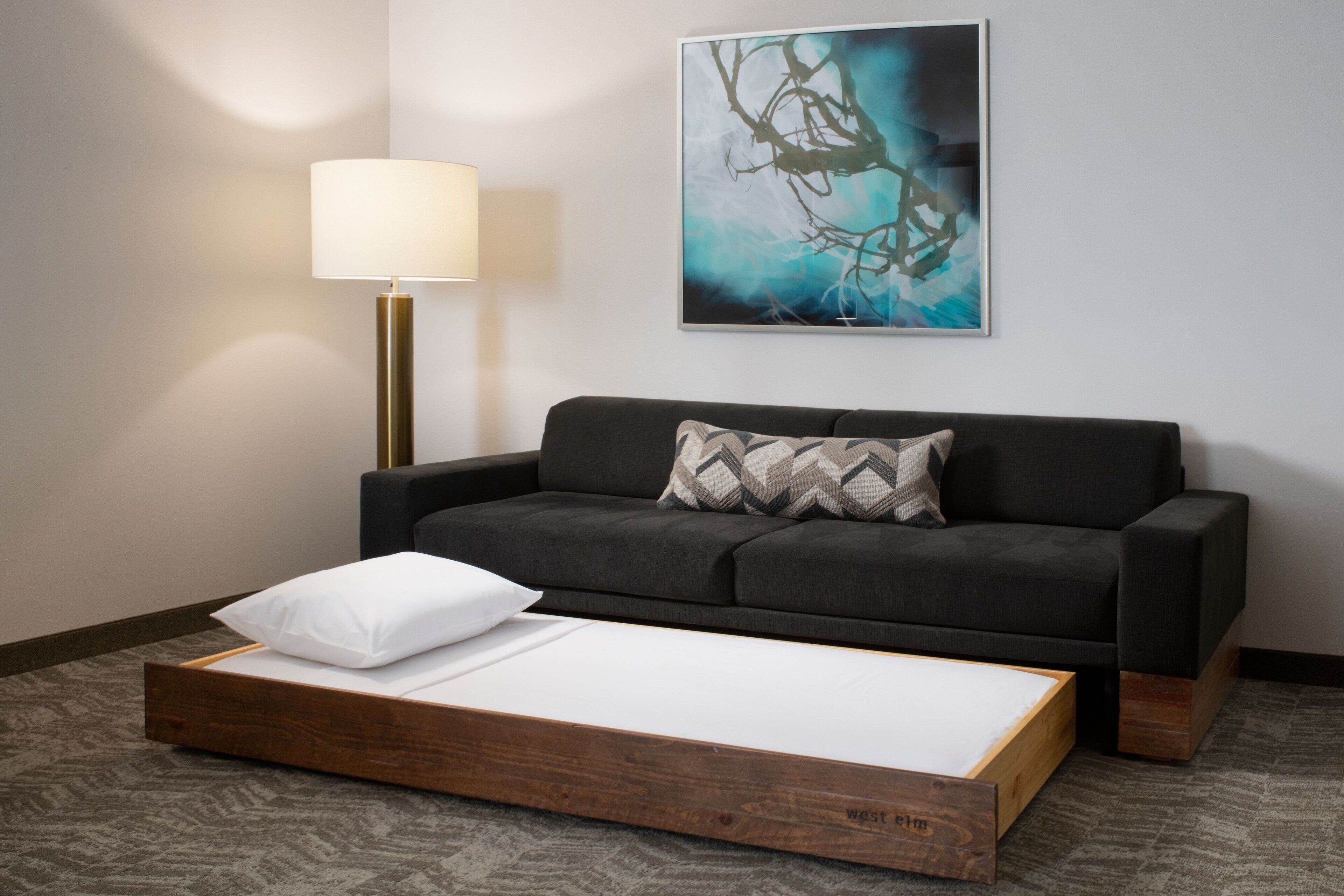 Una habitación moderna con un sofá gris oscuro, una cama plataforma de madera, una lámpara de pie y un cuadro abstracto grande en la pared.