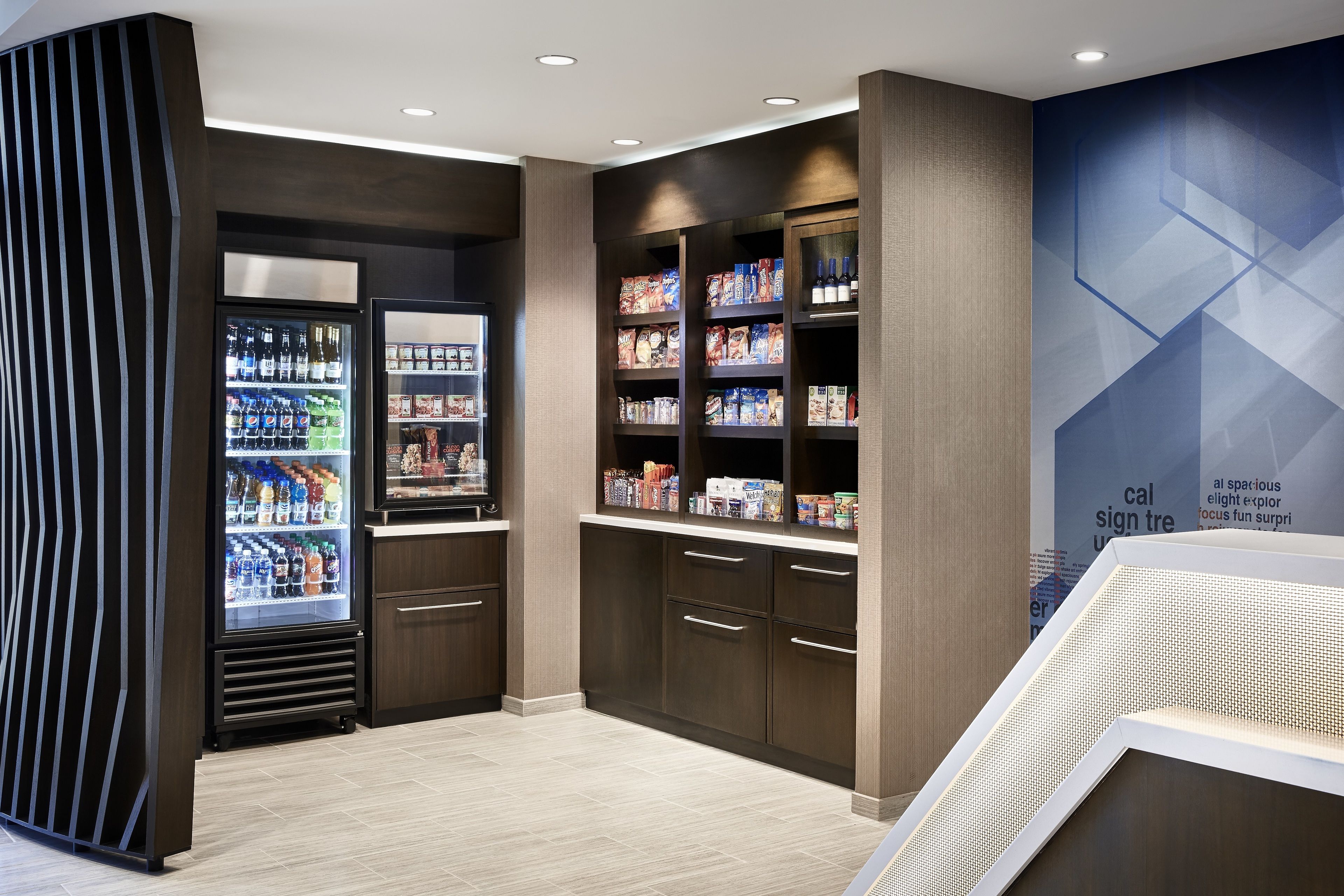 A modern retail space with a refrigerated beverage display, shelves stocked with snacks and packaged goods, and a geometric wall design.
