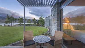 A patio with wicker chairs and a table, overlooking a landscaped garden and a lake, with a stone building and a bed visible through the window. - Mt Michael Lodge (Lowburn)
