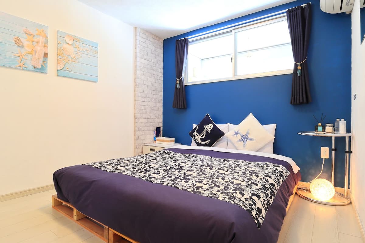 Une chambre à coucher avec un lit, une fenêtre avec des rideaux, une table de chevet et une étagère fixée au mur, sur laquelle sont posés divers objets.