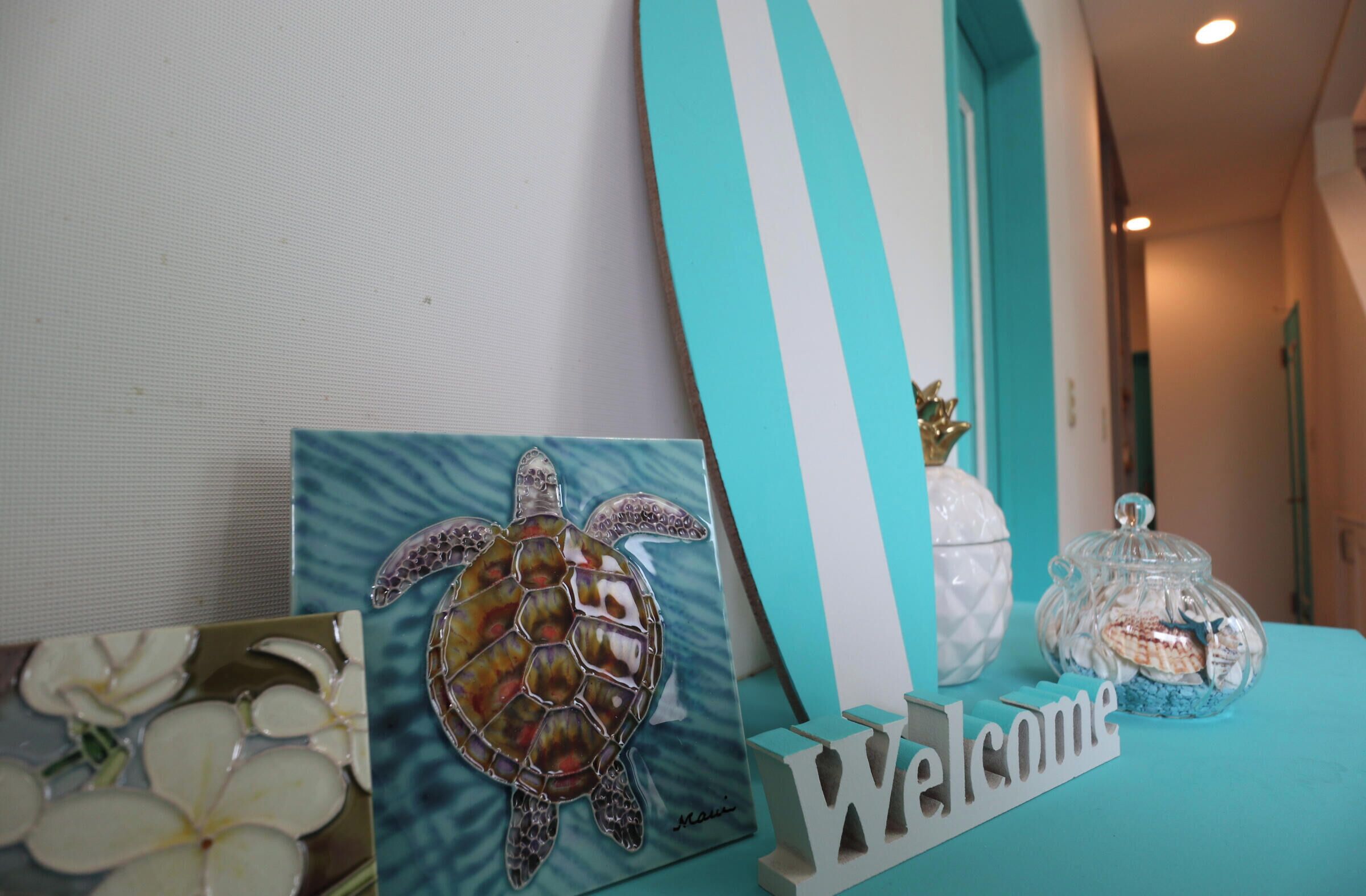 A welcome sign, a turtle decoration, and a jar with a lid on a teal-colored table.