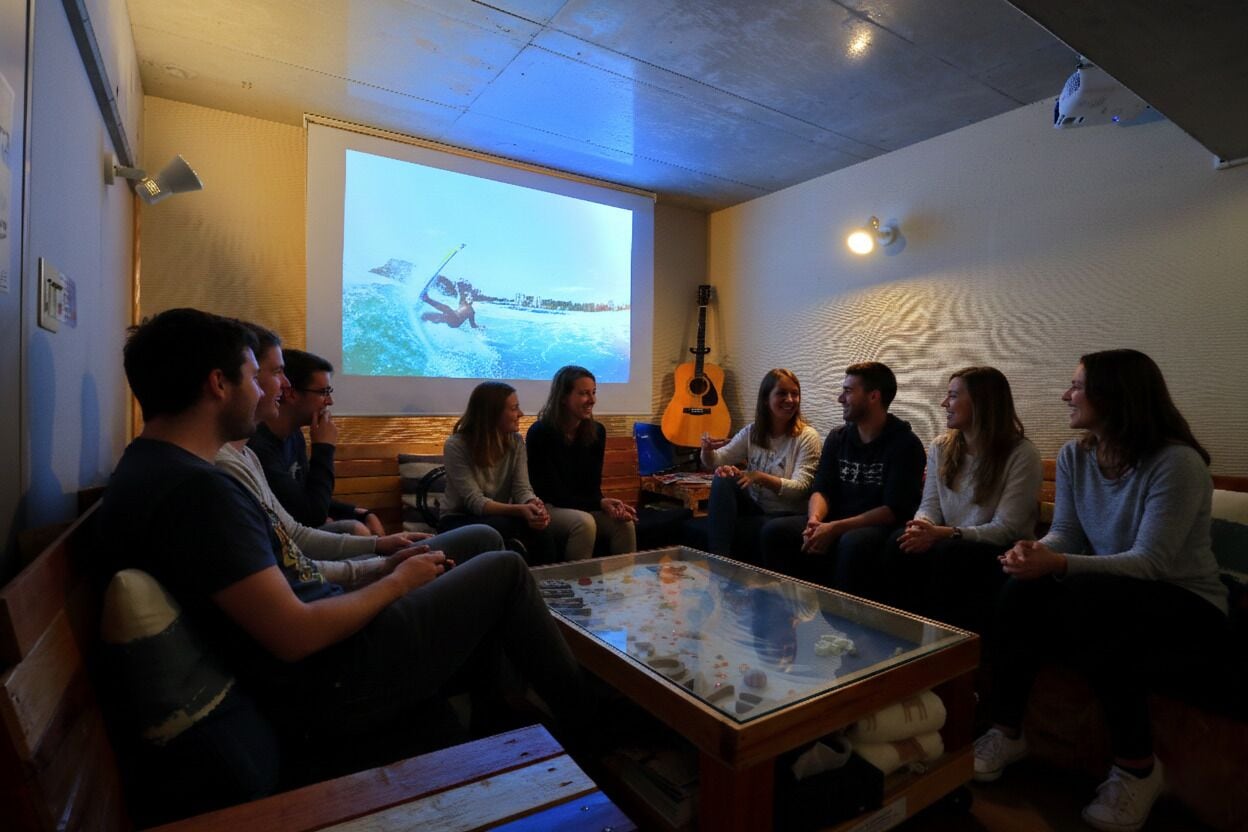 A group of people seated around a glass-top table in a room with a projector screen displaying a video, a guitar, and a textured wall.