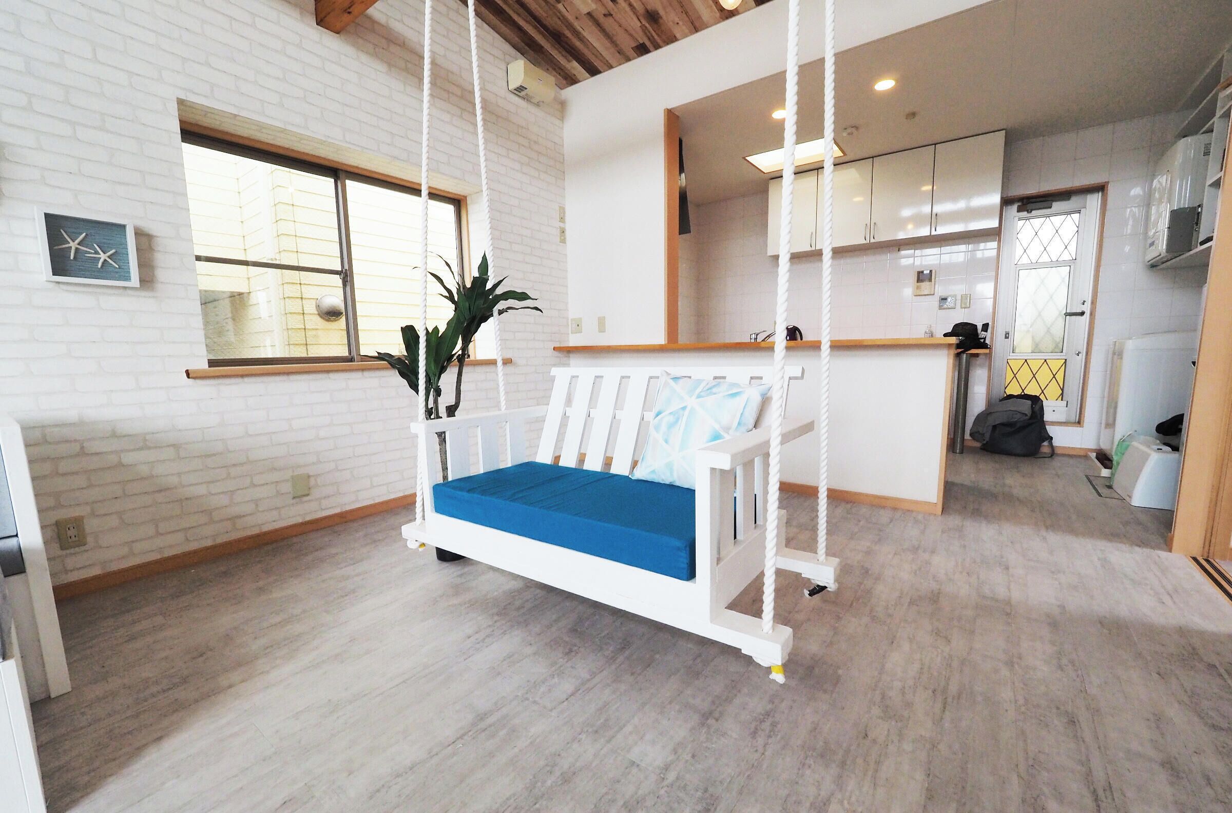A room with a white brick wall, a swing chair with blue cushions, a potted plant, and a kitchen area with white cabinets.