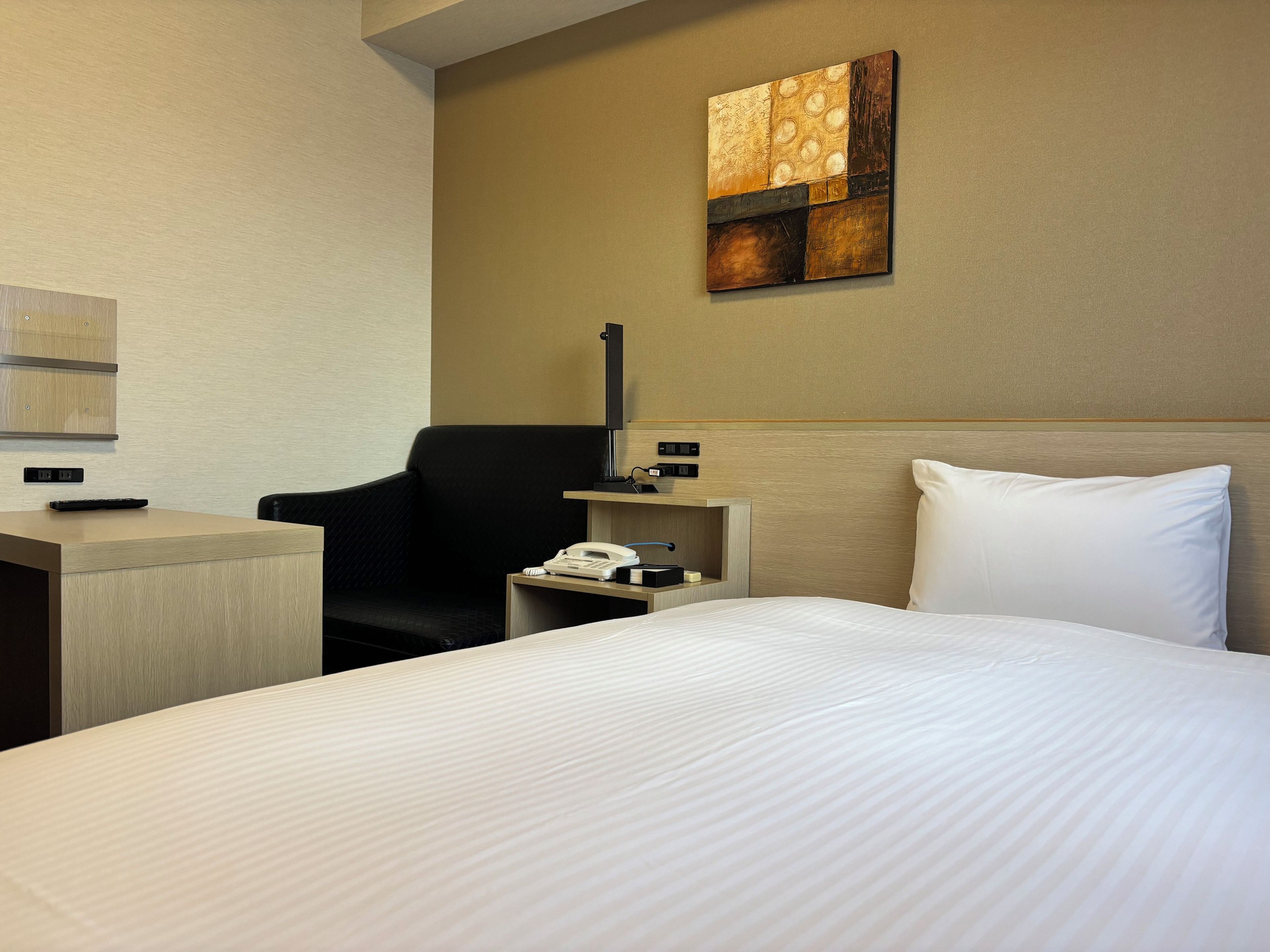 A hotel room with a bed, a desk, a chair, a nightstand, a telephone, and a wall-mounted artwork.