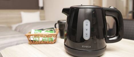 A black electric kettle on a wooden table with a basket of green tea bags and a black mug in the background.