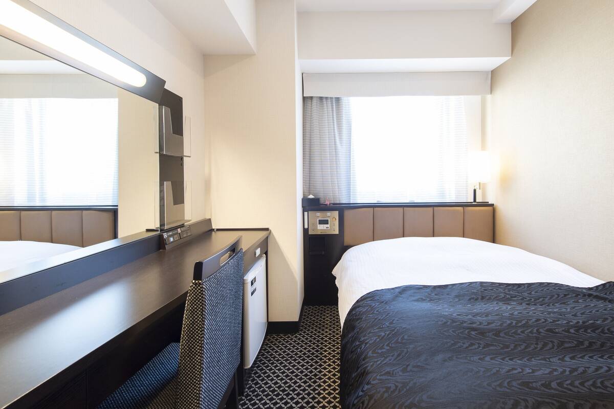 A hotel room with a bed, a desk with a chair, a mirror, a window with curtains, and a bedside table with a lamp.