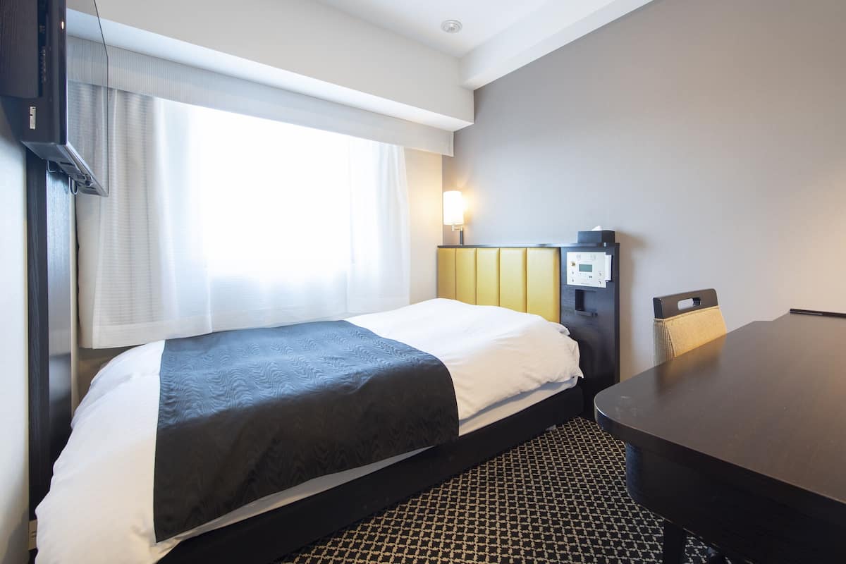 A hotel room with a bed, a desk, a chair, a control panel, and a window with curtains.