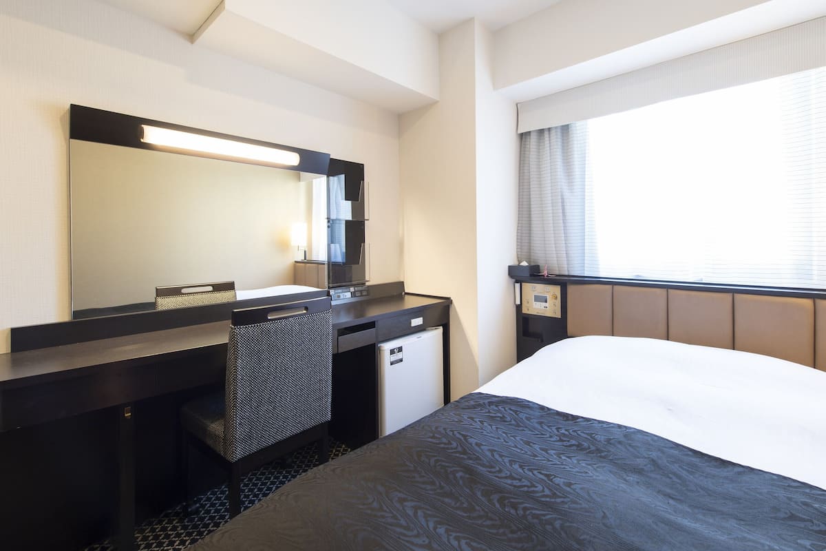 A hotel room with a large bed, a desk with a mirror, a small kitchenette, and a window with curtains.