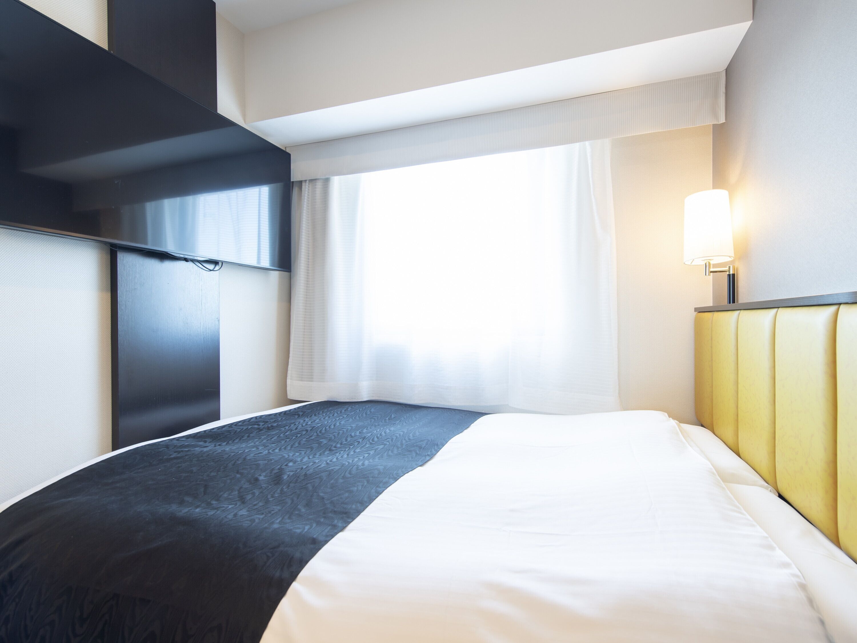 Una habitación de hotel moderna con una cama grande, un televisor de pantalla plana, una lámpara de noche y un ventanal con cortinas.