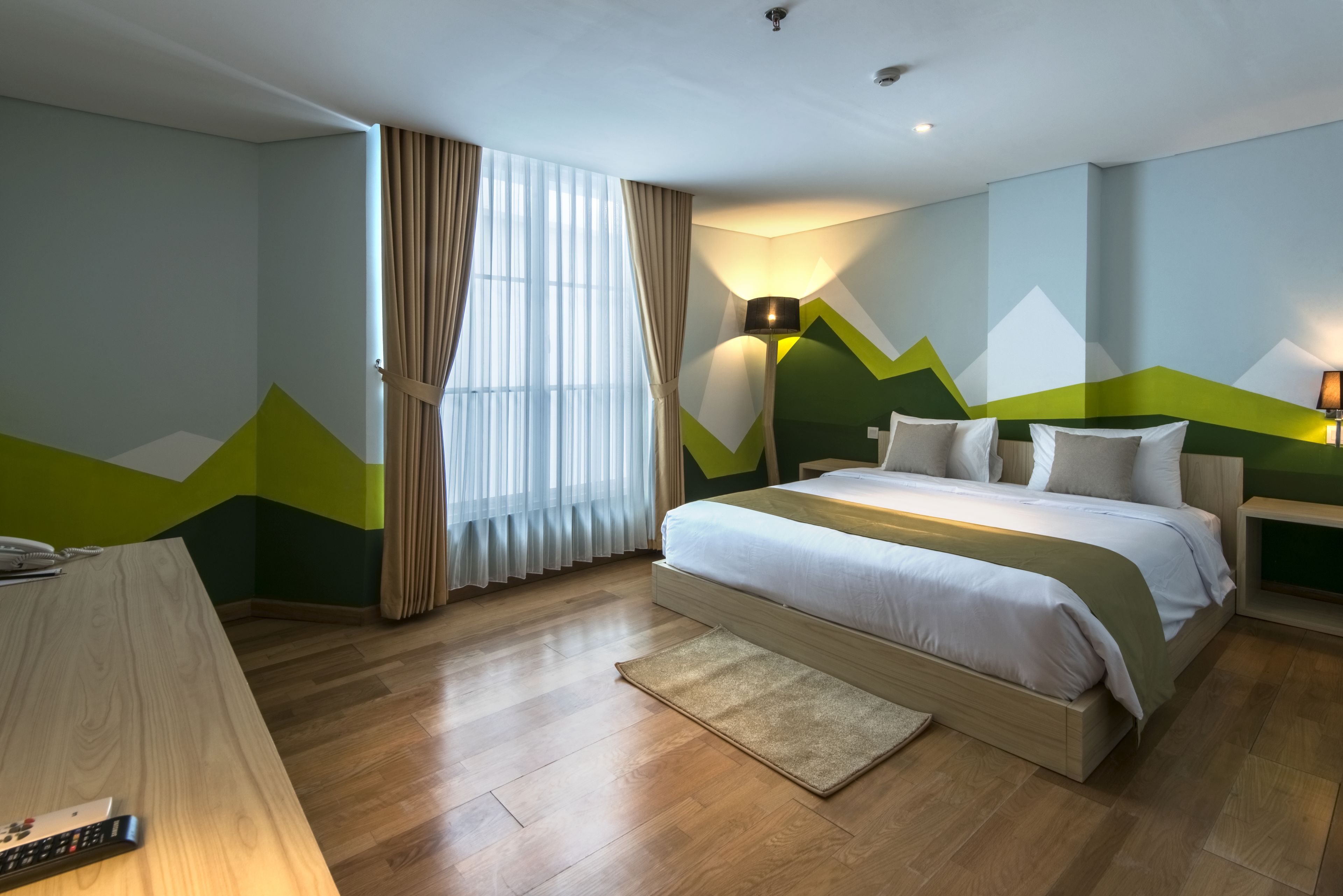 A modern hotel room with a wooden floor, a large bed with white bedding, a desk with a lamp, and a view of the outdoors through sheer curtains.