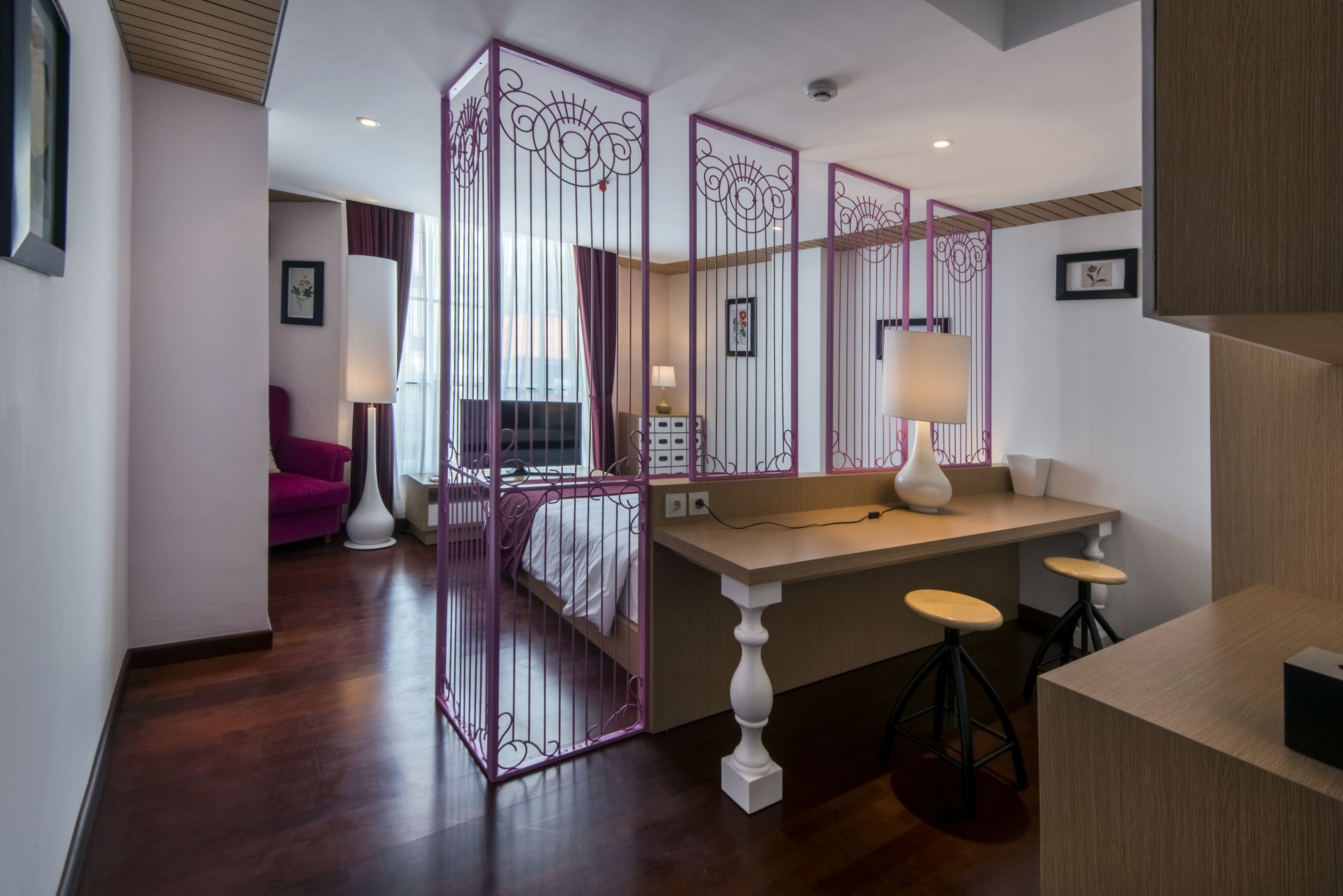 A modern hotel room with a wooden floor, a bed with white bedding, a desk with two stools, and a purple partition.