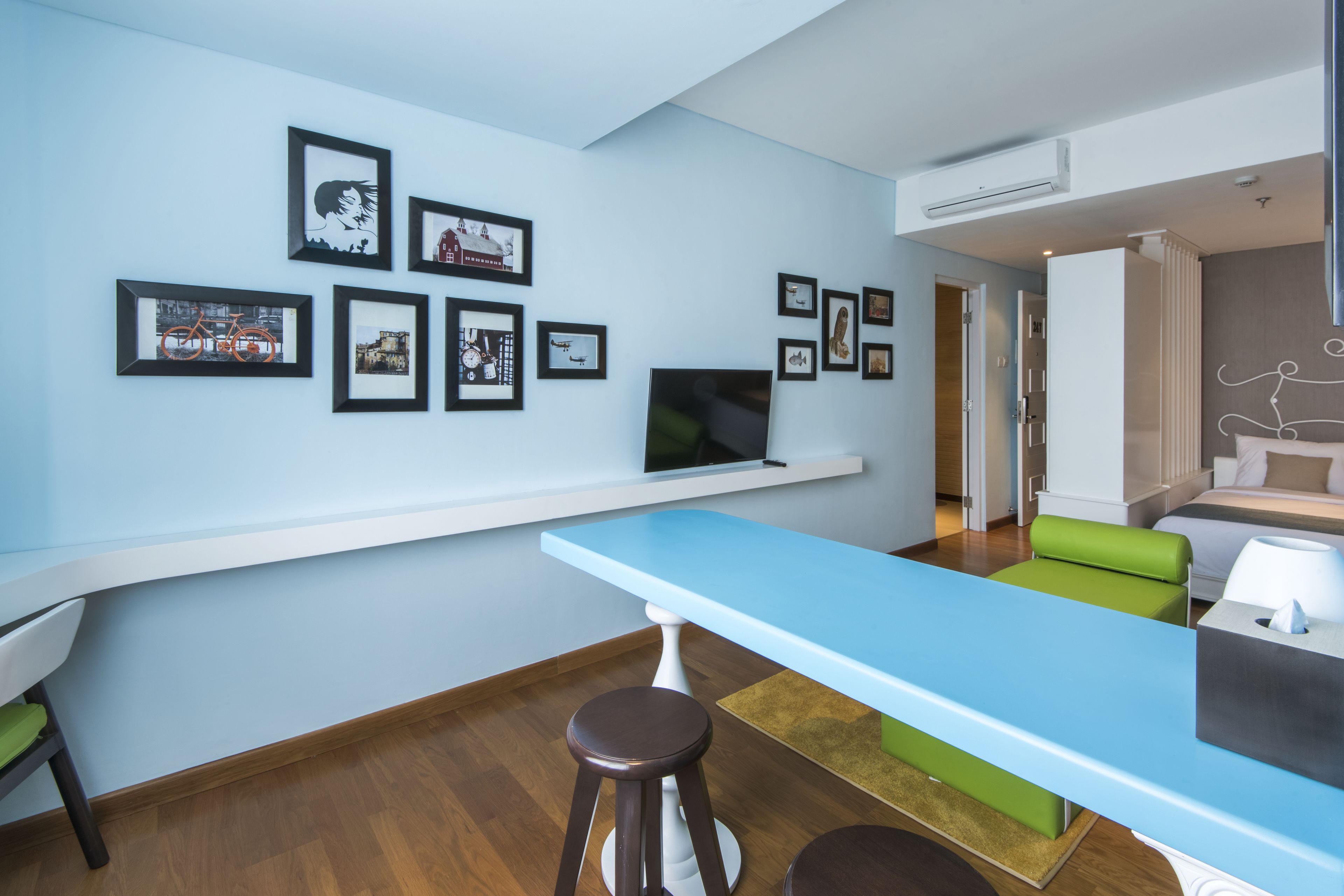 A modern hotel room with a long dining table, a TV, and a seating area with green and white furniture.
