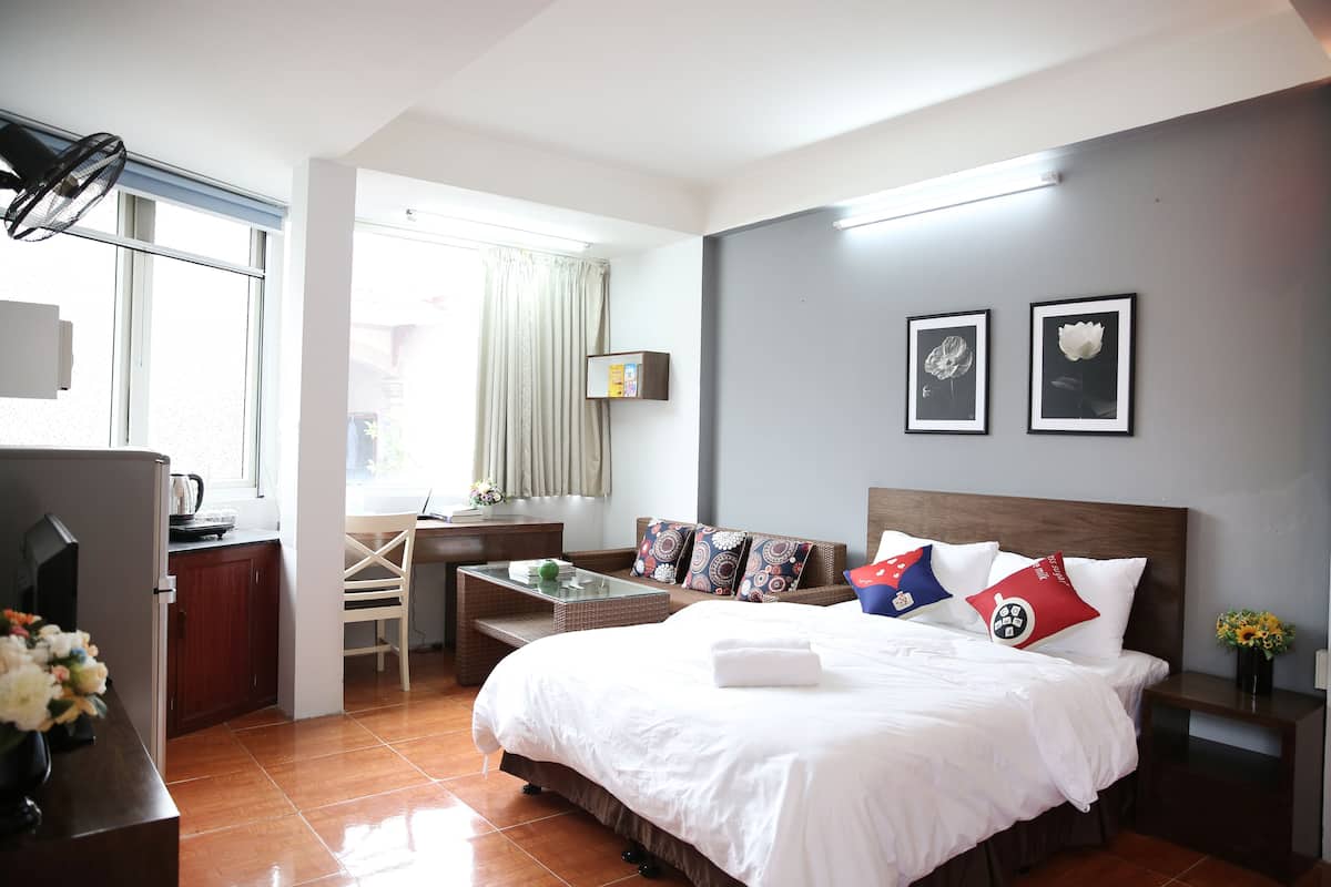 Habitación de hotel con cama, escritorio y silla. Tiene una ventana con cortinas, un televisor en la pared y una mesita con un jarrón de flores.