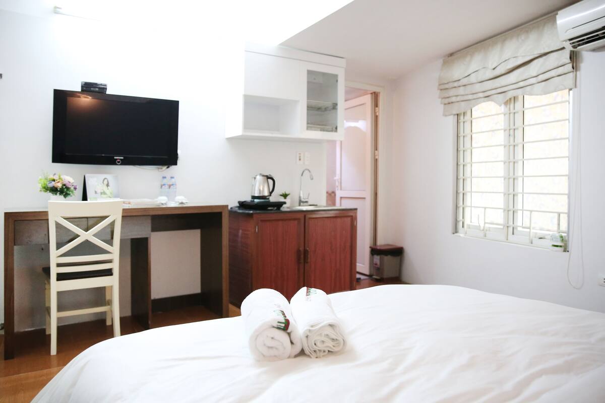 Habitación de hotel con cama, escritorio con televisor, kitchenette y ventana con cortinas.