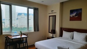 A hotel room with a bed, a desk with chairs, a window with curtains, a nightstand, and a painting on the wall.