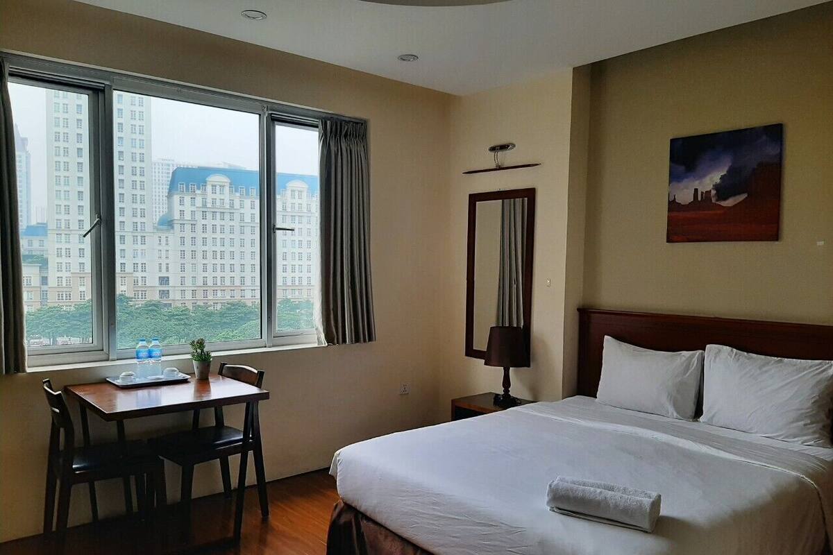 Habitación de hotel con una cama, un escritorio con sillas, una ventana con cortinas, una mesita de noche y un cuadro en la pared.