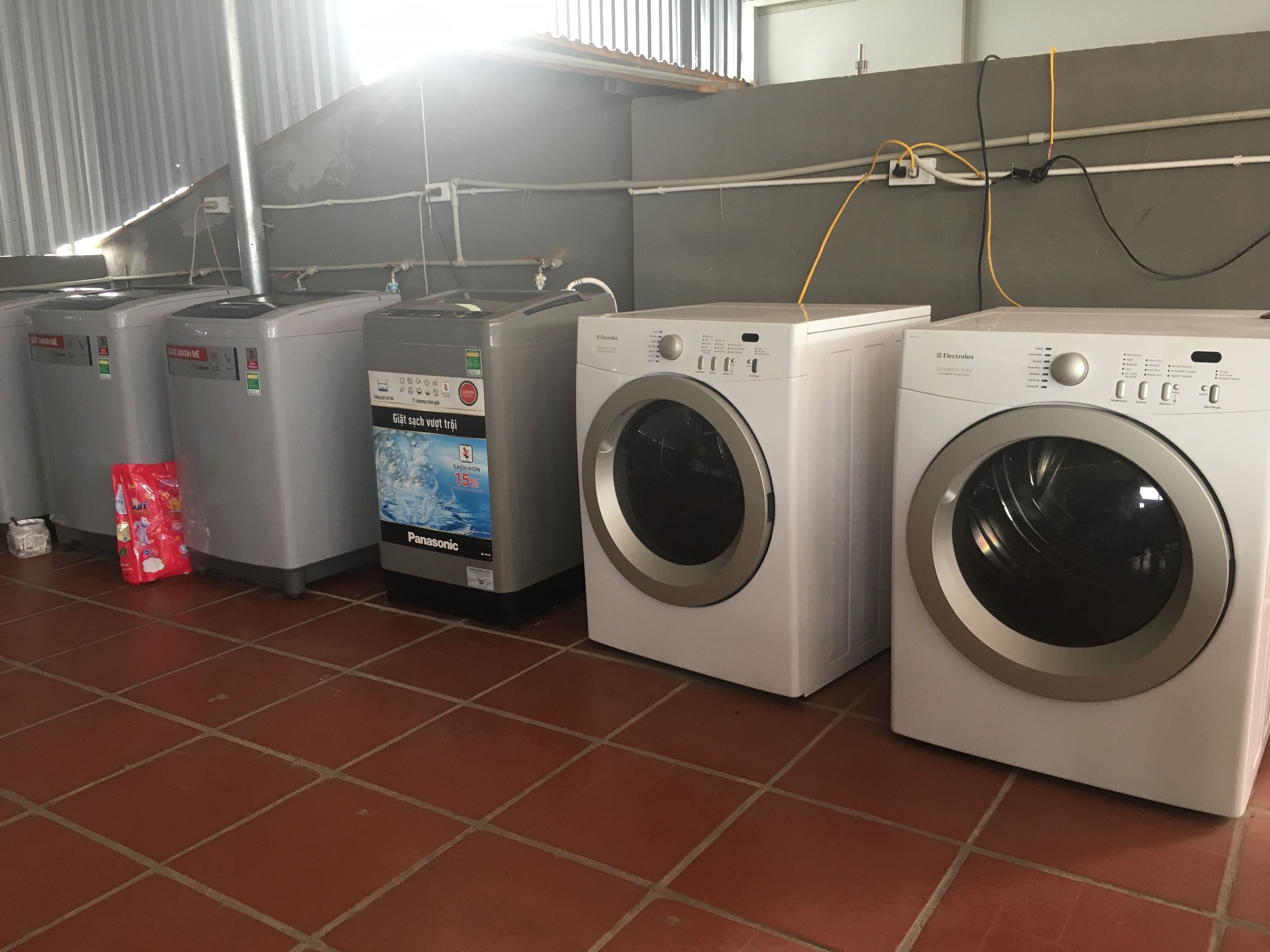 A laundry facility with multiple washing machines and dryers, a red bag of detergent, and visible electrical wiring on the wall.