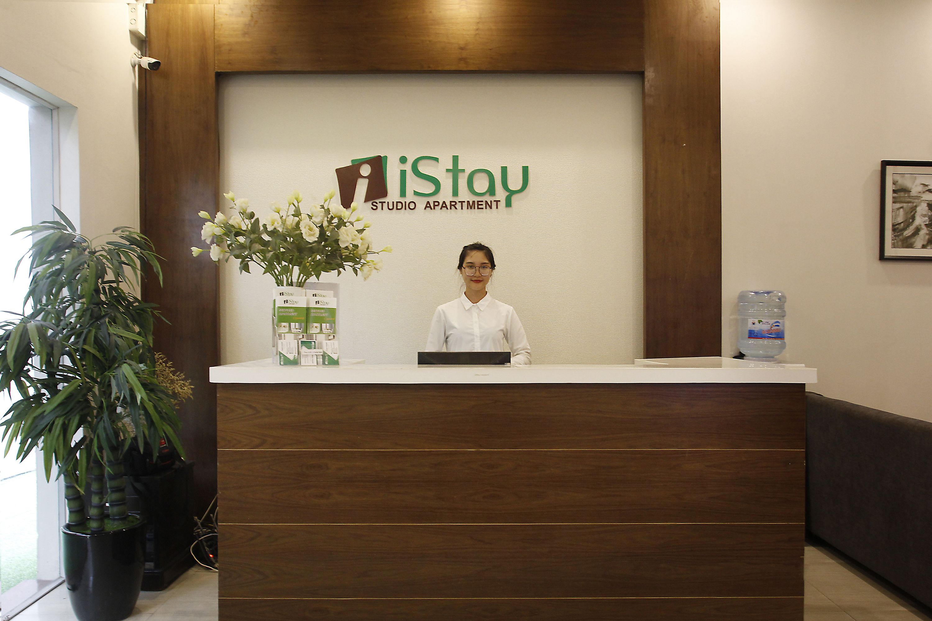 Un comptoir d’accueil se trouve dans l’iStay Studio Apartment, avec un réceptionniste, une plante en pot et un distributeur d’eau.