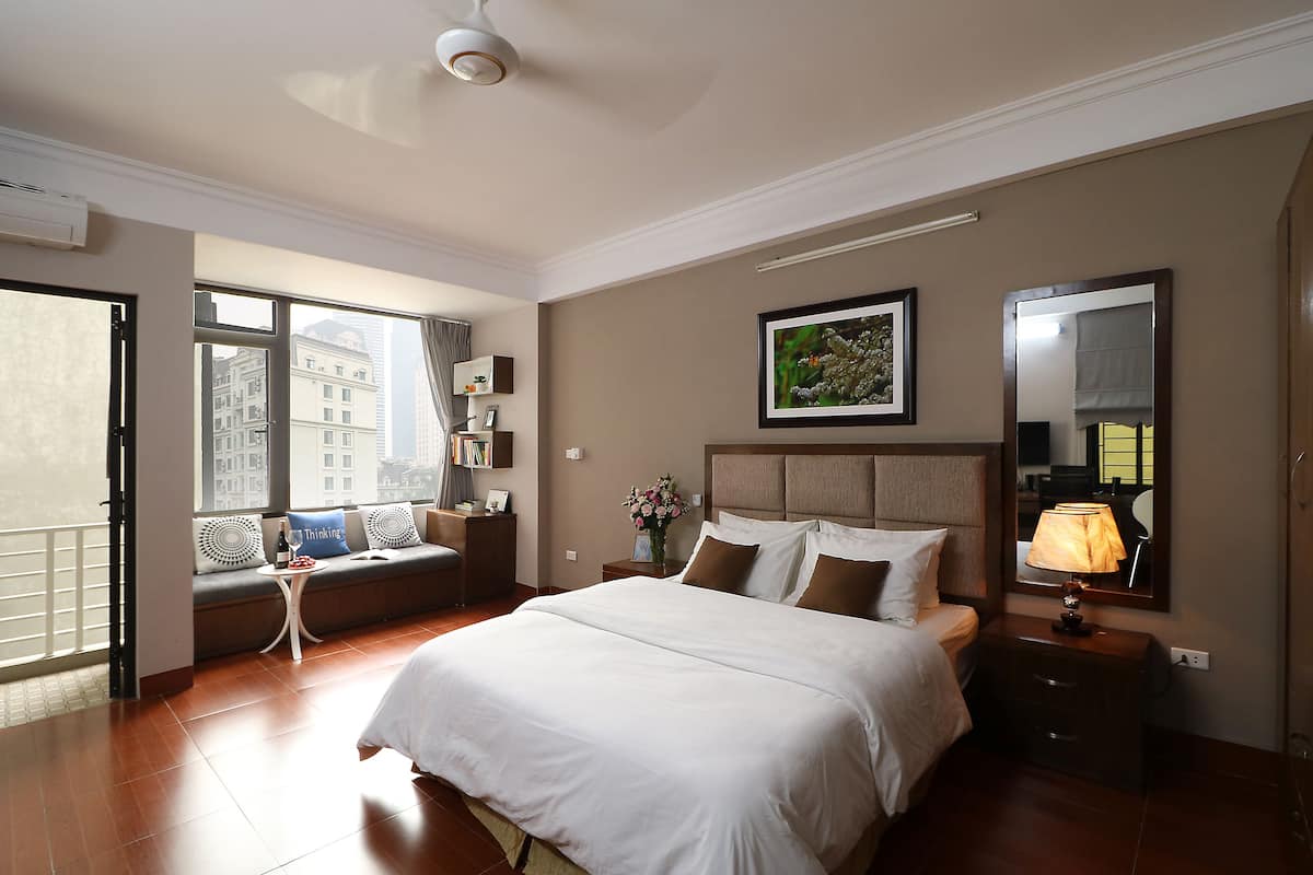 Habitación de hotel con una cama grande, una mesita de noche con lámpara, una ventana con vista a la ciudad y un sofá con almohadones decorativos.