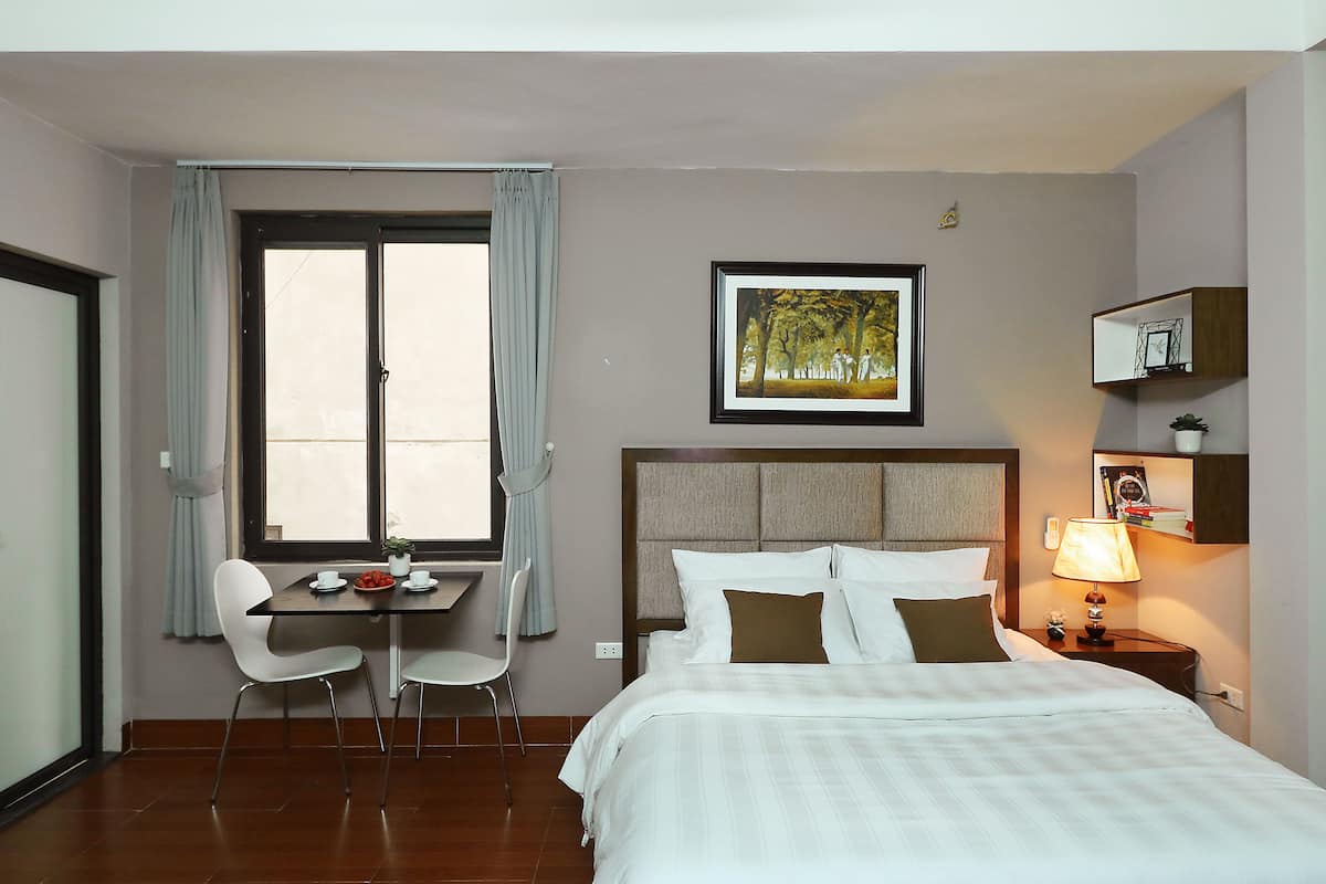 Una habitación de hotel con una cama, un escritorio con sillas, una ventana con cortinas, un cuadro en la pared y una mesita de noche con una lámpara.