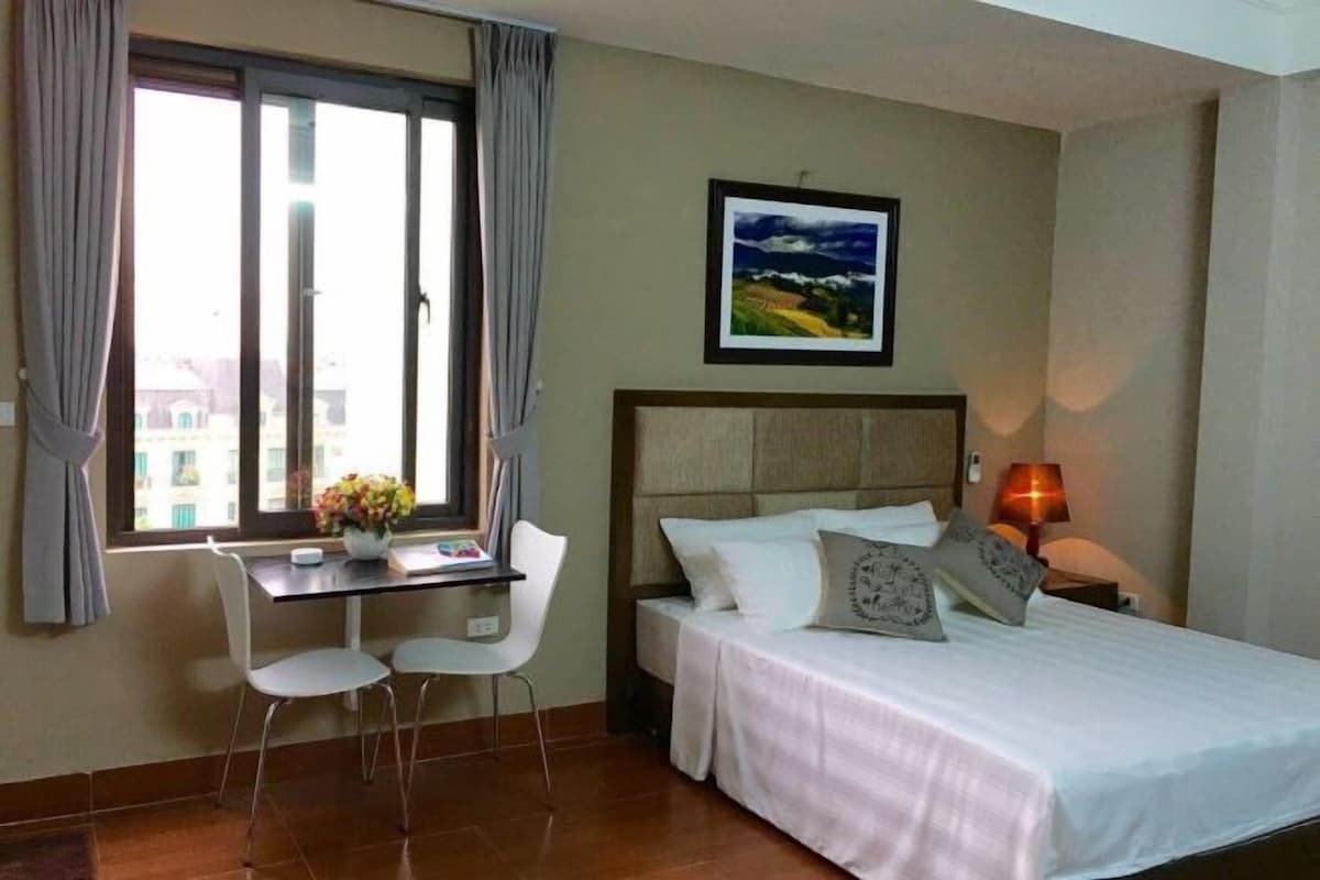 A hotel room with a bed, a desk with two chairs, a window with curtains, a framed picture on the wall, and a bedside lamp.