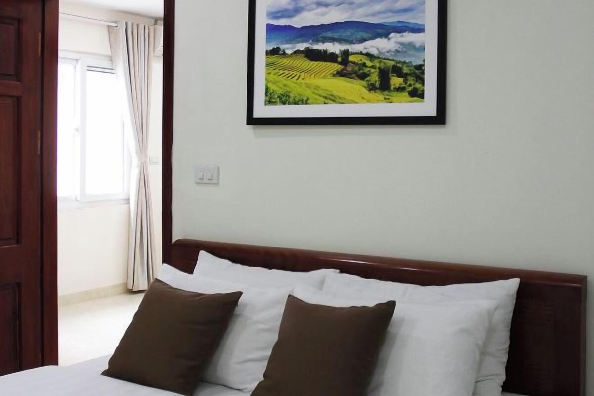 A hotel room with a bed, two pillows, a framed landscape painting, and a window with curtains.