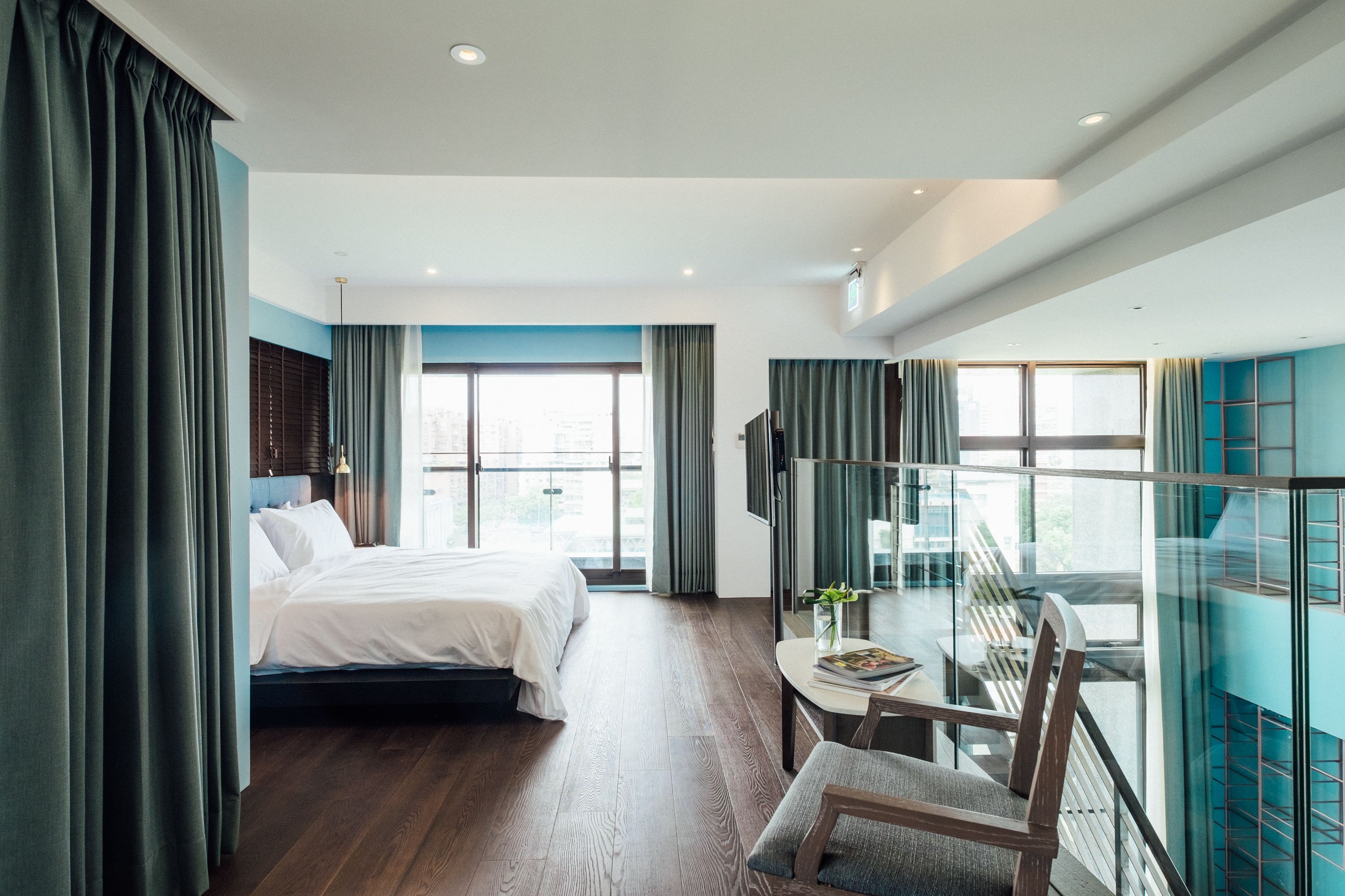 A modern hotel room with a large bed, a desk with a chair, and a balcony with a glass railing.