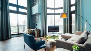 A modern living room with a blue armchair, a grey sofa, and a TV. There are large windows and a city view.