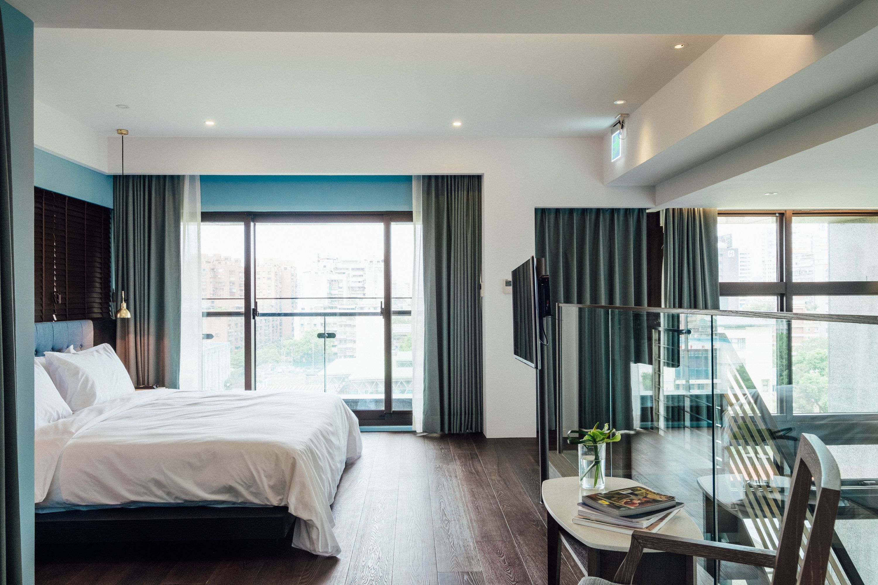 Ein modernes Hotelzimmer mit einem großen Bett, einem Glastisch mit einem Stuhl, einem Fernseher und Blick auf die Stadt durch große Fenster.