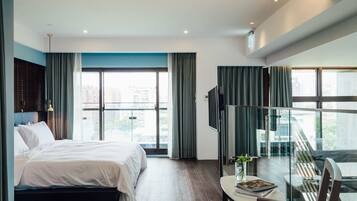 Ein modernes Hotelzimmer mit einem großen Bett, einem Glastisch mit einem Stuhl, einem Fernseher und Blick auf die Stadt durch große Fenster.