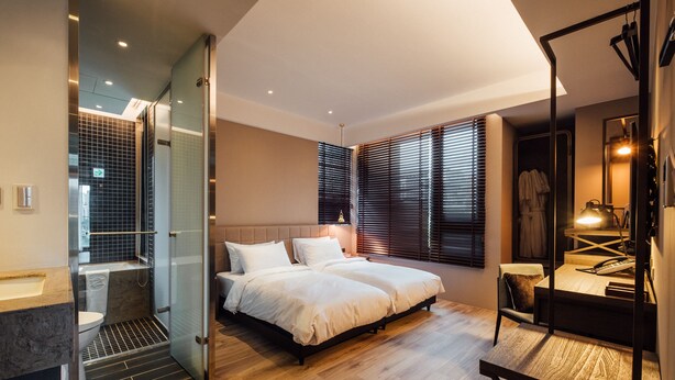 A modern hotel room with a glass-enclosed shower, a large bed with white linens, a wooden desk, and a mirror.