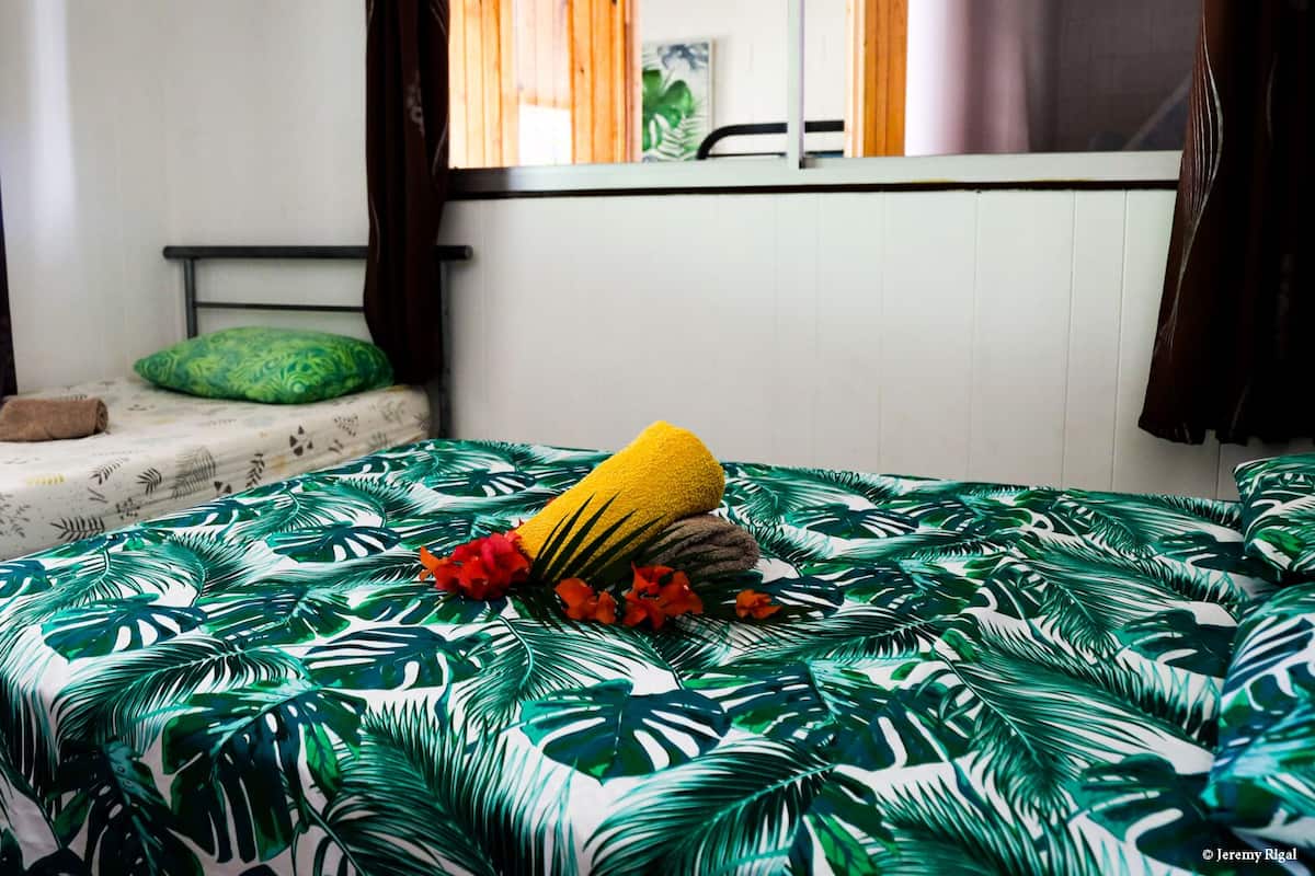 A room with a bed covered in a tropical leaf pattern, a small bedside table with a green pillow, and a window with curtains.