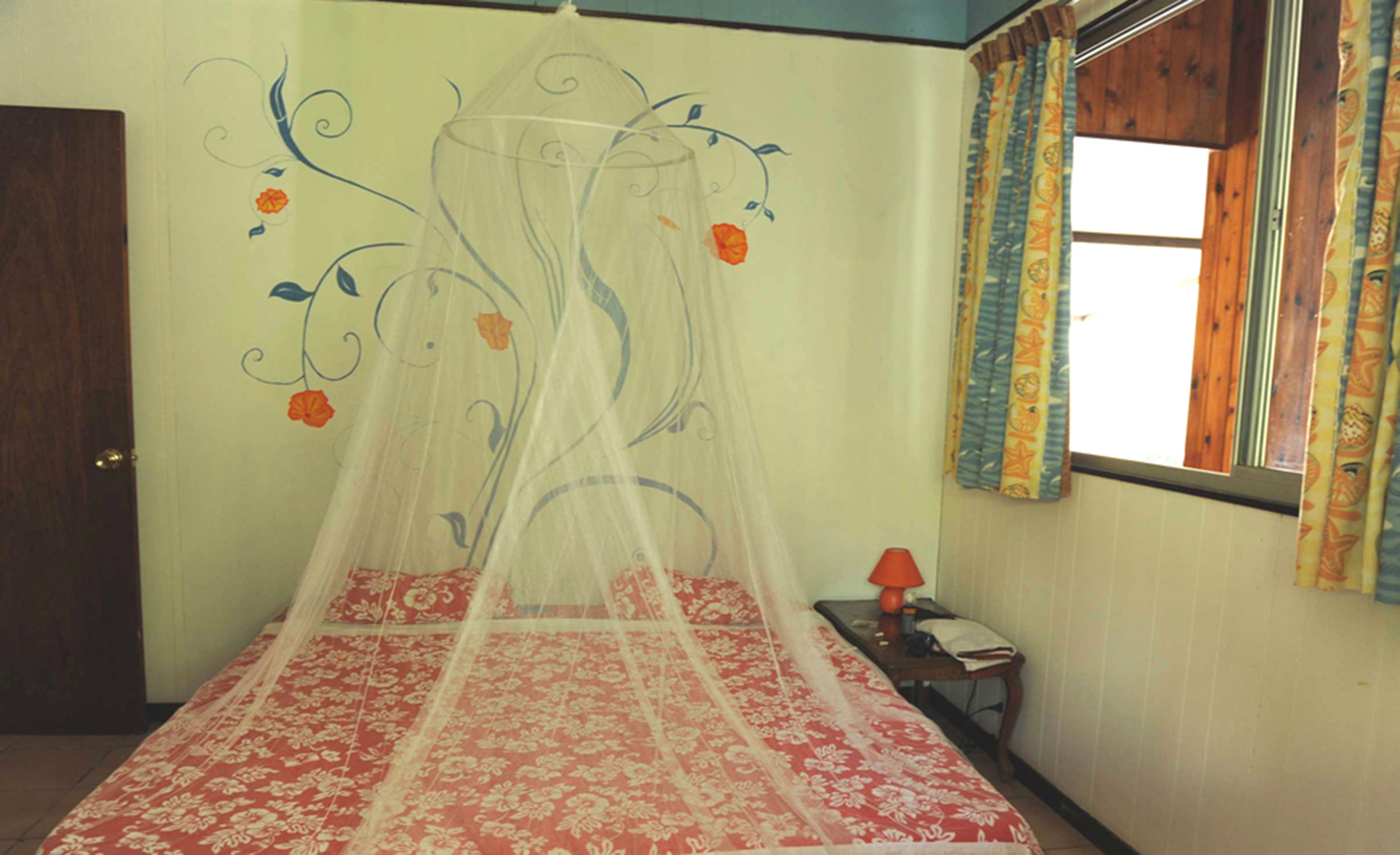 A room with a bed covered by a canopy with floral patterns, a small bedside table with a lamp, and a window with curtains.