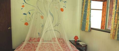 A room with a bed covered by a canopy with floral patterns, a small bedside table with a lamp, and a window with curtains.