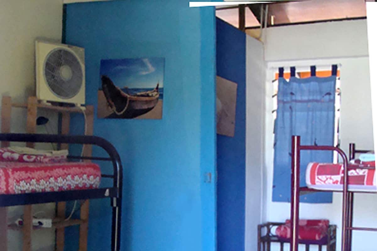 A dormitory room with bunk beds, a fan, a painting, and a desk with a chair.