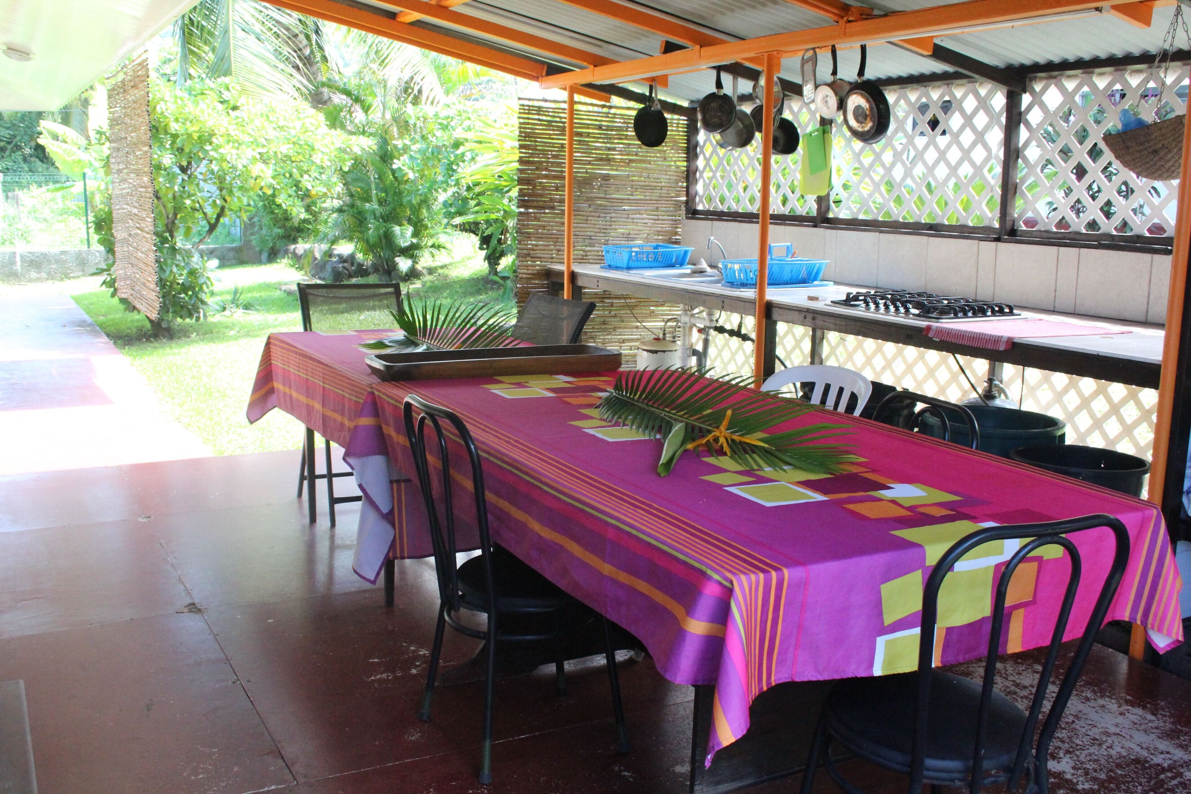An outdoor dining area with a table covered in a colorful tablecloth, surrounded by chairs. There are hanging pots and pans above the table, and a fence with a lattice design in the background.