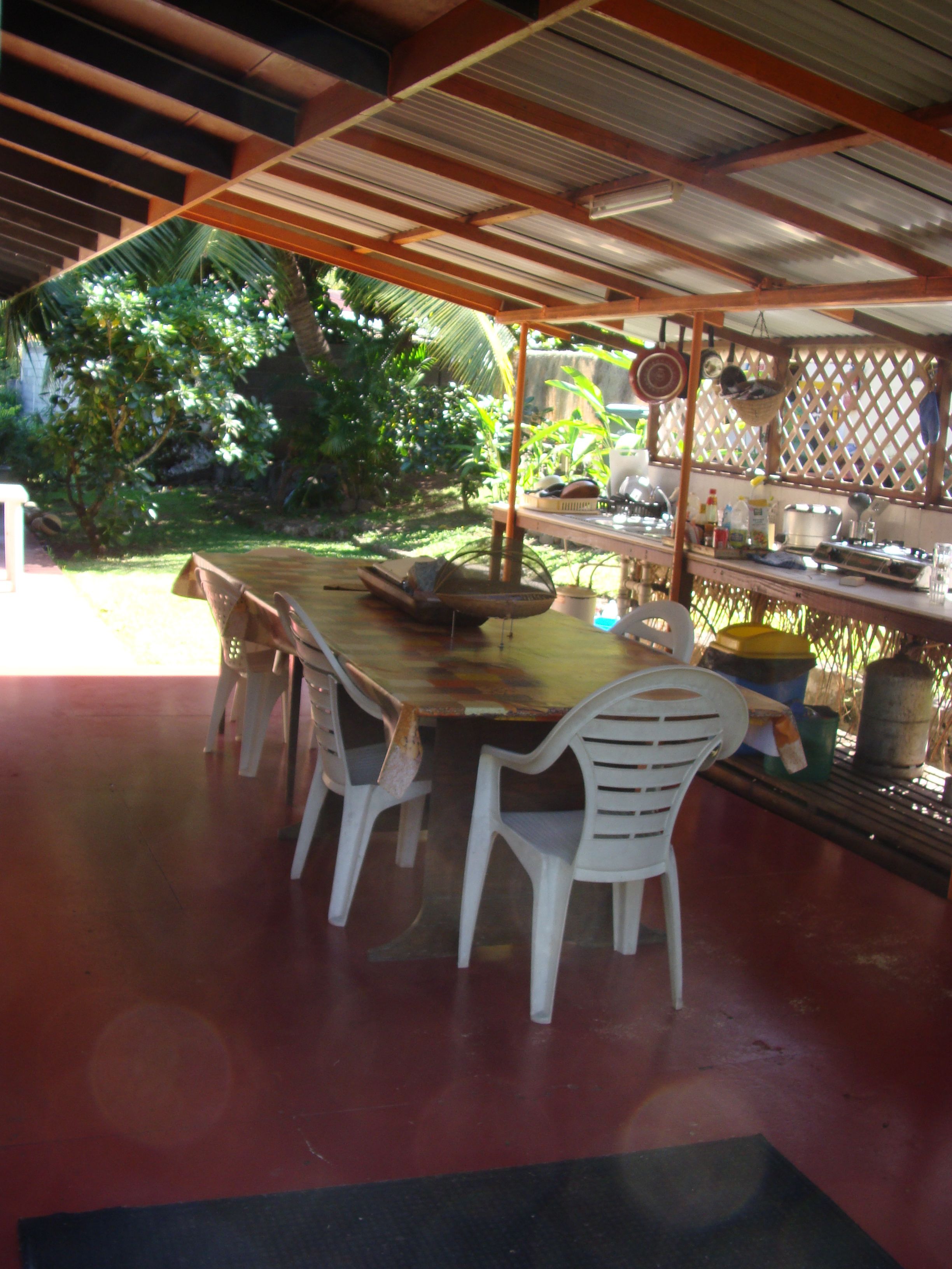 An outdoor dining area with a wooden table and white plastic chairs, surrounded by lush greenery.
