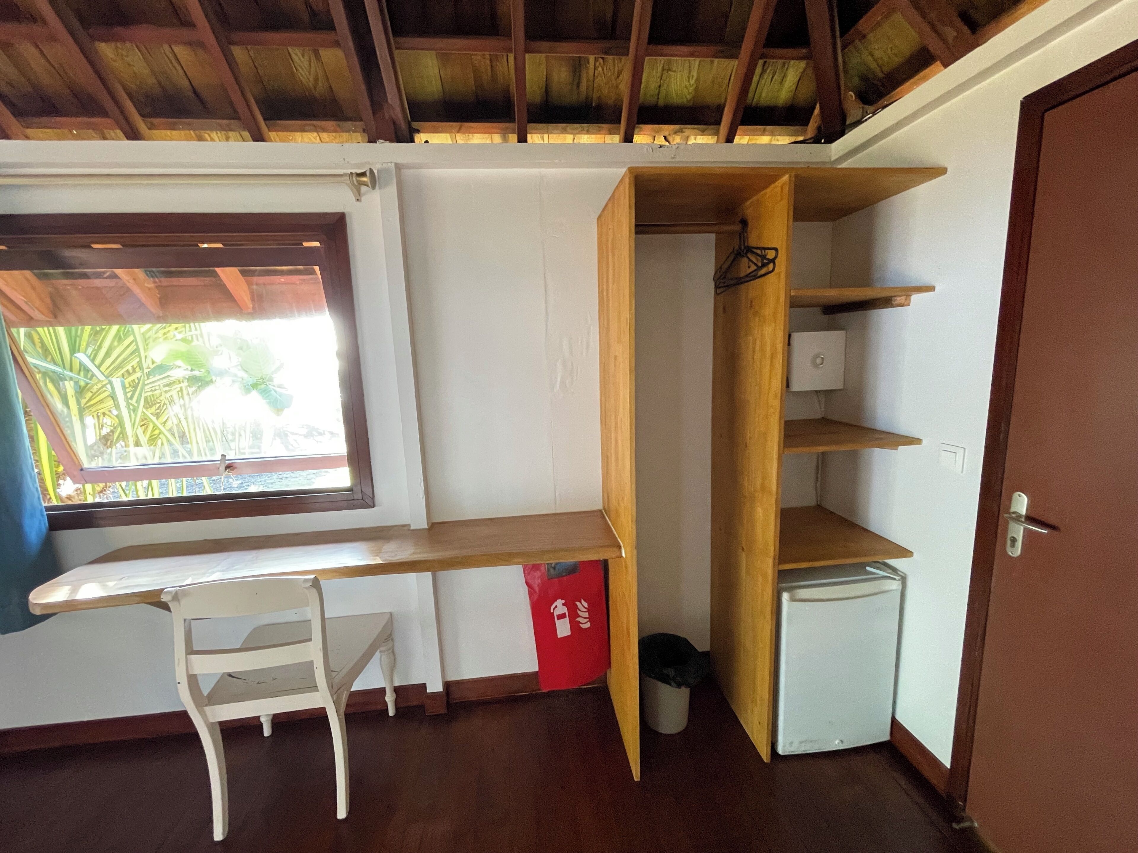 Una habitación con un escritorio de madera, una silla, un refrigerador blanco pequeño y un bote de basura rojo.