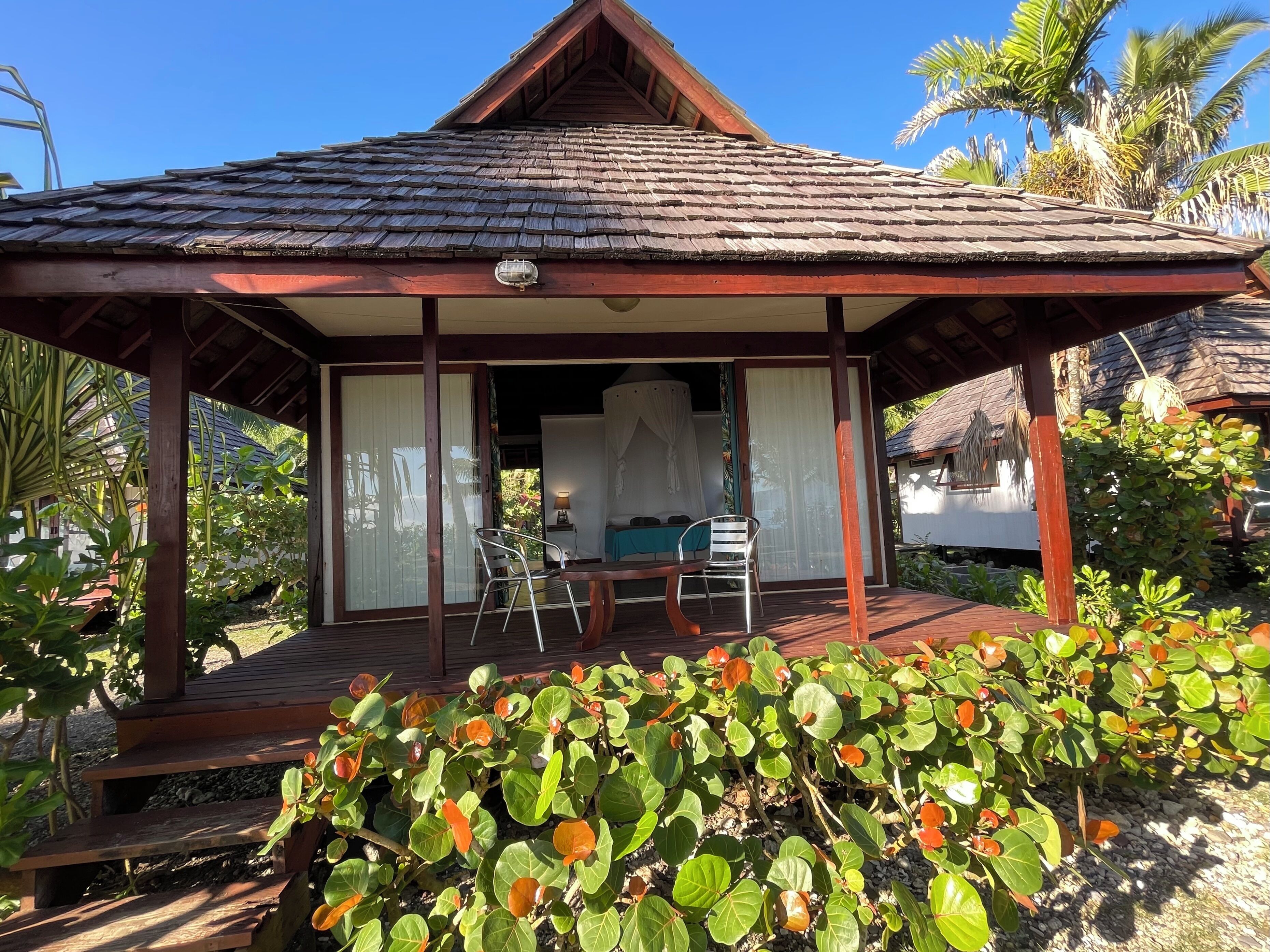 A wooden gazebo with a tiled roof, a covered seating area with a table and chairs, and a view of a lush garden with tropical plants.