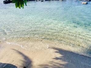 A clear beach with visible sand and shallow water, a shadow of a tree, and rocks in the background. - Punatea Village (Taiarapu-Est)