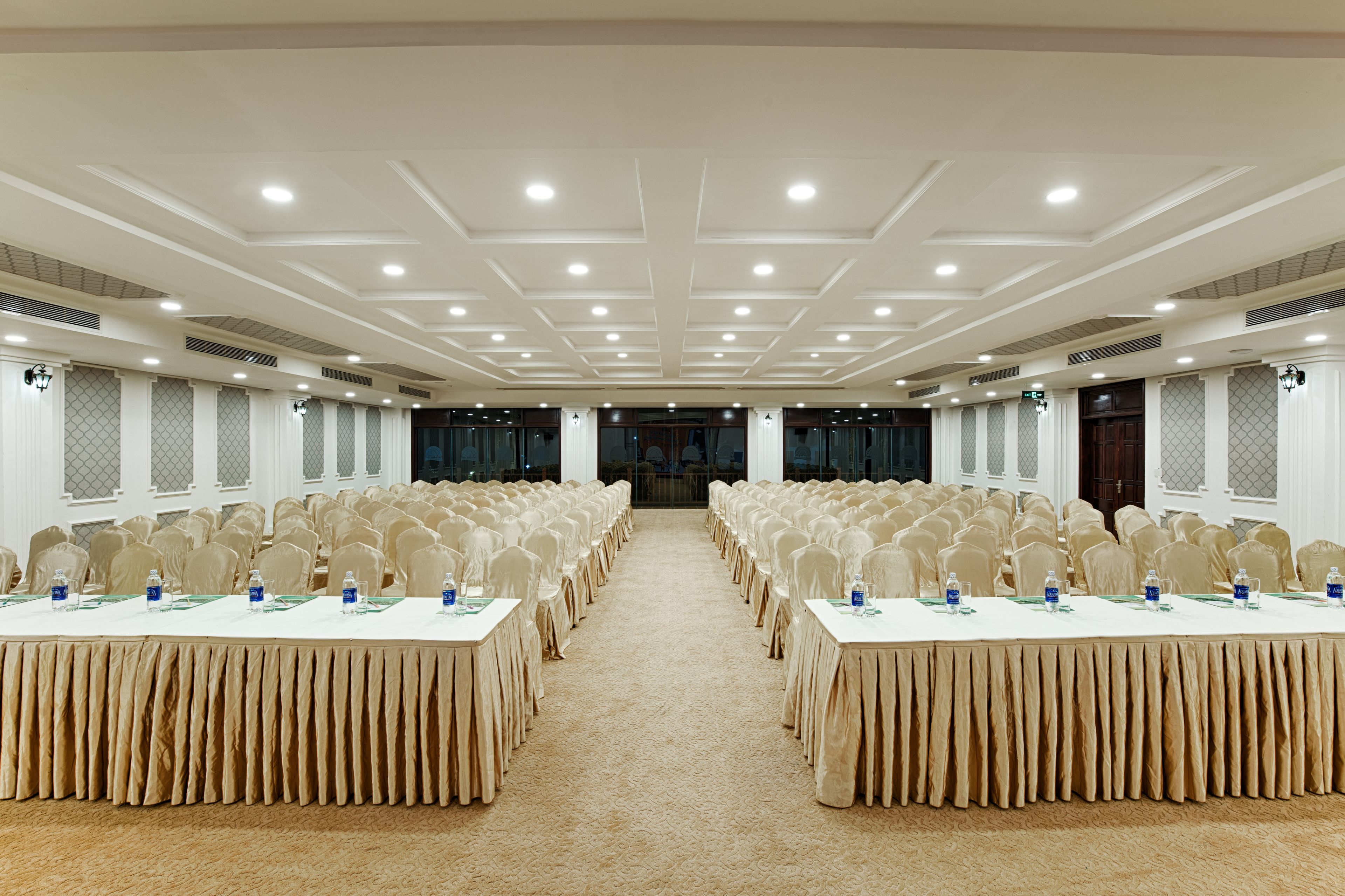 A conference room with rows of tables and chairs, a carpeted floor, and a high ceiling with recessed lighting.