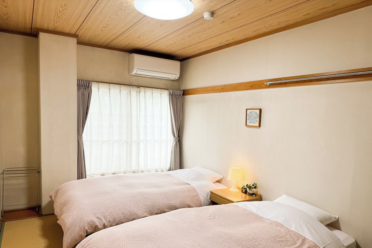 A hotel room with two beds, a bedside table with a lamp, an air conditioning unit, and a window with curtains.